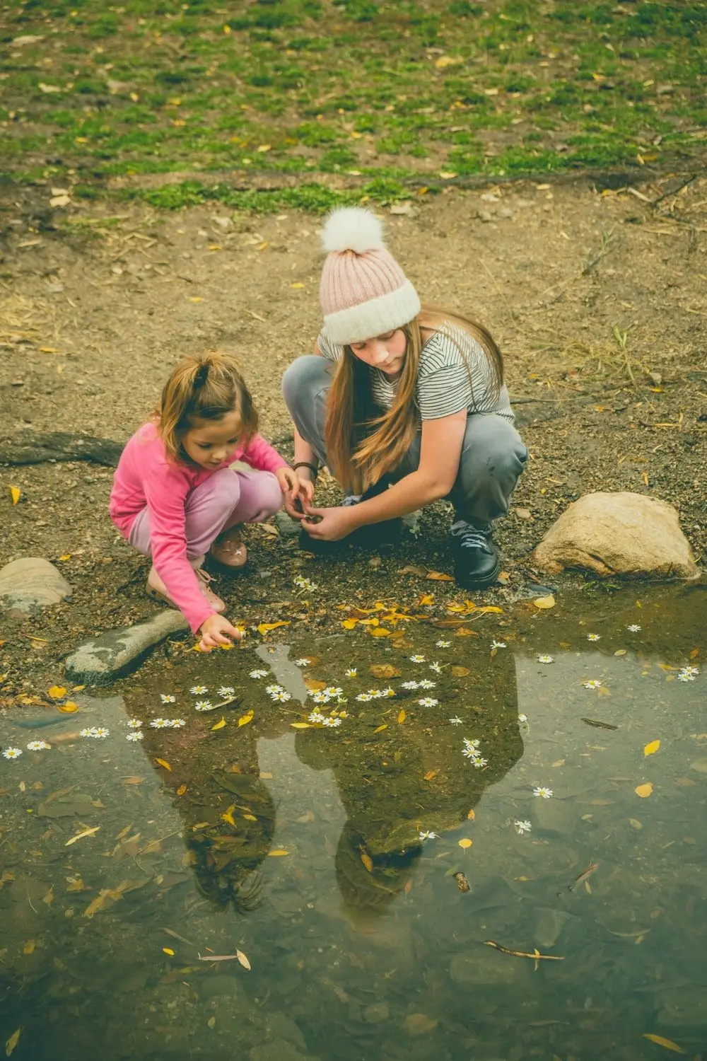 Two children crouching at the edge of a pond, exploring nature together among floating daisies.